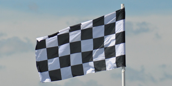 Symbolbild einer schwarz-weiß gemusterten Zielflagge, die im Wind weht. Im Hintergrund ist ein bewölkter Himmel zu sehen.