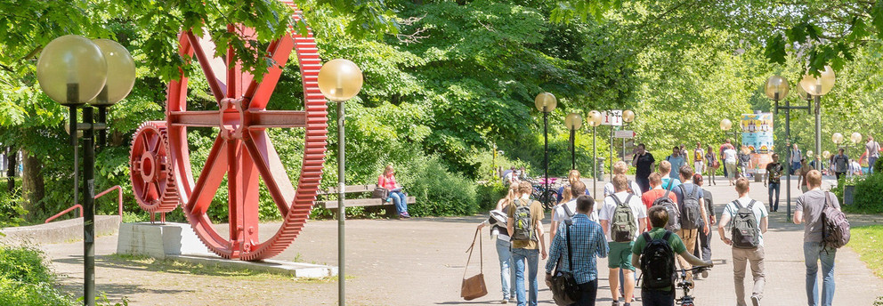 Studierende laufen über den Campus Nord. Es ist eine große rote Skulptur in Form von großen Zahnrädern zu erkennen.