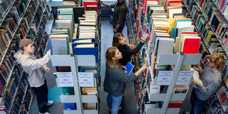 Studierende stehen suchend an mehreren Bücherregalen in der Bibliothek.