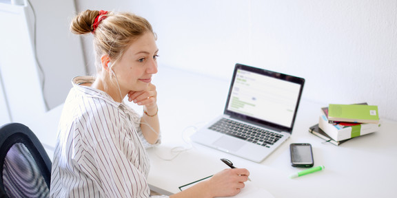 Frau sitzt mit Laptop am Schreibtisch und notiert etwas in ein Notizbuch